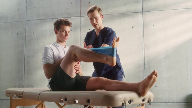 Sport Physiotherapy Specialist Showing How to Stretch a Rubber Band on Specific Muscle Groups or Joints to Young Male Athlete. Sportsman Recovering from Mild Injury, Undergoing Rehabilitation.