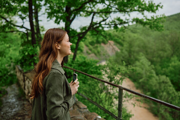 Naklejka na ściany i meble a woman in a raincoat takes pictures on the phone of a beautiful view of the forest. Travel, technology, mobile photography