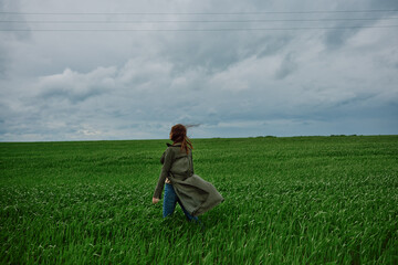 Naklejka na ściany i meble a woman in a long raincoat stands in a green field in windy weather with her back to the camera