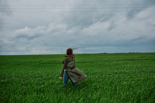 a woman in a long raincoat stands in a green field in windy weather with her back to the camera