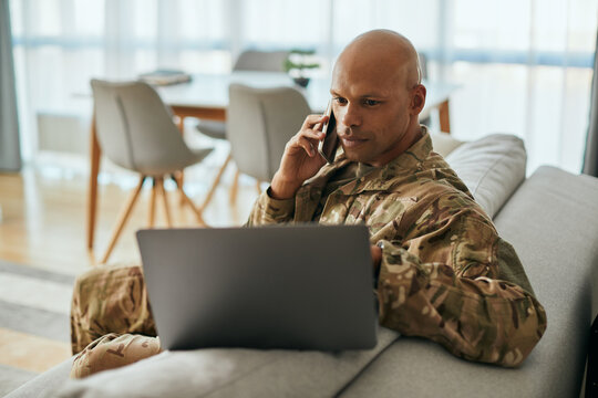 Young black soldier talks on cell phone while surfing the net on laptop at home.