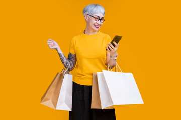 Asian happy senior female woman tattoo holds colourful shopping packages standing on yellow...