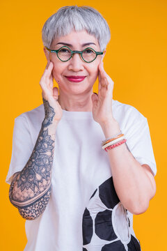 Cheerful Smiling Asian Senior Woman Wear Glasses Grey Hair In Casual Cloth Hand Tattoo Abstract Pattern Portrait Headshot On Yellow Color Background