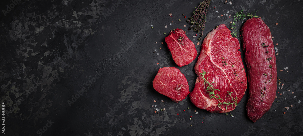 Wall mural Assortment of raw cuts of raw beef meat steaks with spices on a dark background. Long banner format. top view