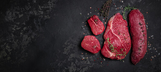 Assortment of raw cuts of raw beef meat steaks with spices on a dark background. Long banner...