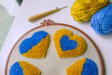 Closeup of bulky tufted hearts on grey fabric in a hoop, punch needle and two balls of yarn in blue...