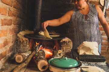 Fototapeta na wymiar Mujer mexicana torteando maza de maíz en un metate y una estufa de leña para hacer tortillas caseras 
