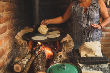 Mujer mexicana torteando maza de maíz en un metate y una estufa de leña para hacer tortillas...