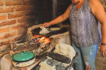 Mujer mexicana torteando maza de maíz en un metate y una estufa de leña para hacer tortillas...