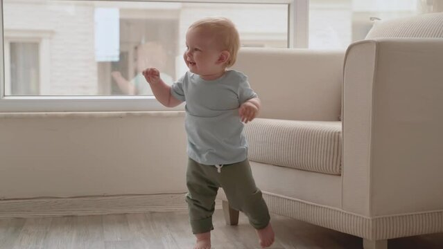 Single adorable baby child learning to walk in light living room. Cheerful laughing toddler boy doing his first steps from one armchair to another. Happy childhood and family concept. Slow Motion