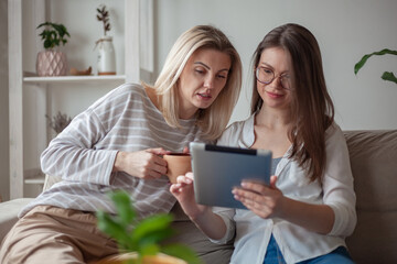 Two young women friends using tablet together, sitting on the couch and smiling