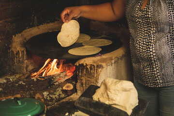 Mujer mexicana torteando maza de maíz en un metate y una estufa de leña para hacer tortillas...