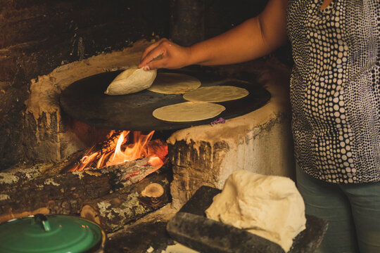 Mujer mexicana torteando maza de maíz en un metate y una estufa de leña para hacer tortillas caseras	