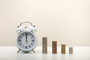 white alarm clock on the white table and a pile of coins , as time passing concept for business deadline, time and money, copy space
