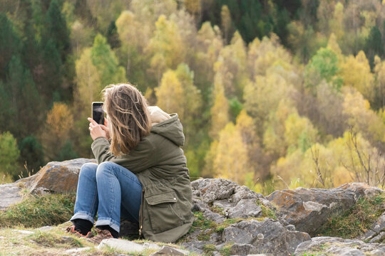 Woman sitting in nature wearing a green jacket and blue jeans, taking a photo of the amazing landscape behind her