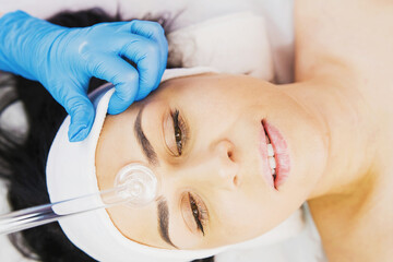 Naklejka premium Closeup photo of professional beautician giving darsonval active electric facial procedure to young woman in beauty salon