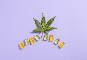 Top view colorful gummy bears and cannabis leaves on light violet background. Copy space.