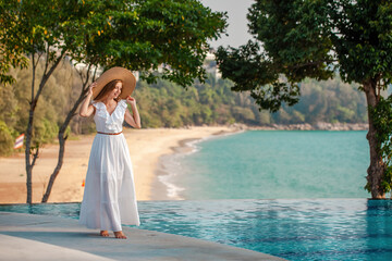 Young travel woman in white dress and hat walking near blue swimming pool with sea view in modern tropical resort on sunset. Summer beach vacation in Thailand