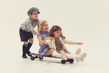 Portrait of three cheerful children playing together, boy rolling girls on skateboard isolated over...