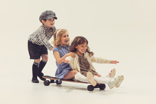 Portrait of three cheerful children playing together, boy rolling girls on skateboard isolated over grey studio background