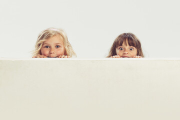 Portrait of two little beautiful girls, children peeking out the table isolated over grey studio...