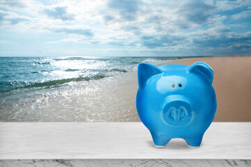 Fototapeta premium Saving money for summer vacation. Piggy bank on white marble surface near sandy beach and sea, space for text