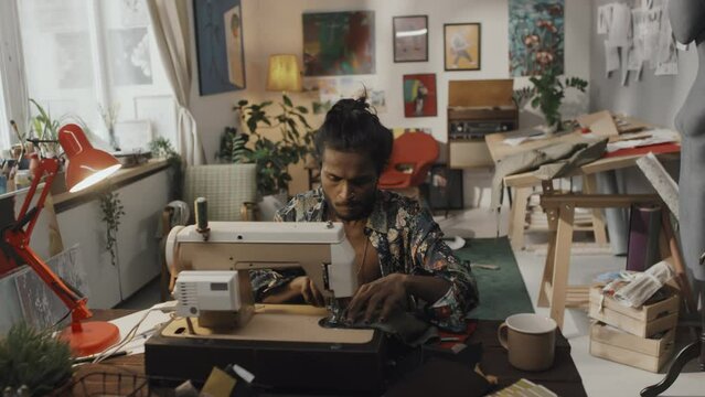 Handsome young Indian fashion designer working on sewing machine in creative retro style messy workshop