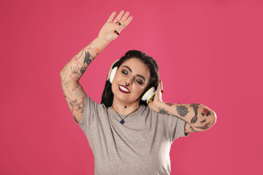 Beautiful young woman with tattoos on arms, nose piercing and dreadlocks listening to music against pink background
