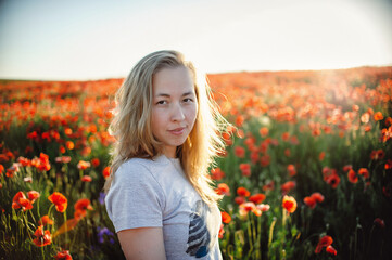 Fototapeta na wymiar Portrait of a beautiful Caucasian woman smiling and looking into the camera lens during sunset. Outdoor portrait of a smiling white girl. Happy cheerful girl laughs in a field of red flowers