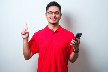 Young handsome Asian wearing casual shirt and glasses holding smartphone and pointing up with fingers number one while smiling confident and happy