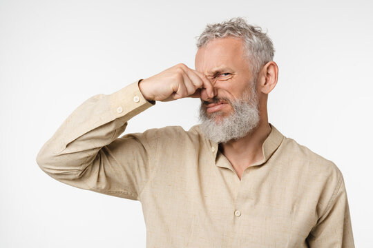 Mature middle-aged caucasian man in beige shirt with grey hair smelling stinky, closing nose with a hand because of odor stench isolated in white background