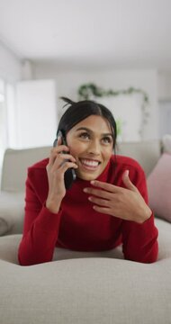 Vertical video of happy biracial woman talking by phone on sofa