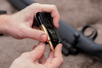 A male hunter equips a rifle magazine with live ammunition against the background of his weapon, shell casings and bullets.
