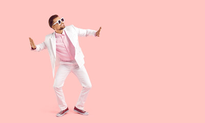 Funny guy dancing on copy space studio background. Happy handsome young man wearing white suit and trendy sunglasses dancing isolated on pastel pink empty copyspace background