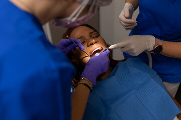 A black woman patient keeps her mouth open while dental hygienist checks and cleans gums and teeth...