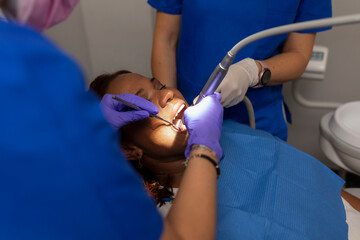 A black woman patient keeps her mouth open while dental hygienist checks and cleans gums and teeth...