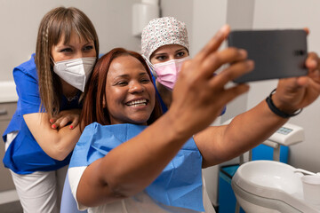 A black woman patient, takes a happy selfie photo with the smartphone side by side the dentist and dental hygienist at the dental clinic - obrazy, fototapety, plakaty