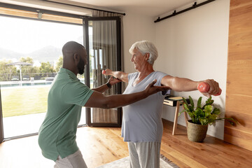 Fototapeta na wymiar African american male physiotherapist helping caucasian senior woman to do exercise with dumbbells