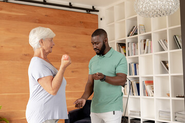 African american male physiotherapist helping caucasian senior woman to do exercise with dumbbells - Powered by Adobe