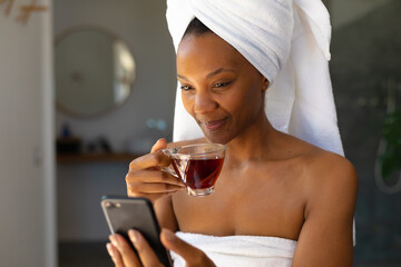 African american woman in a bathrobe holding a tea cup using smartphone at home - obrazy, fototapety, plakaty