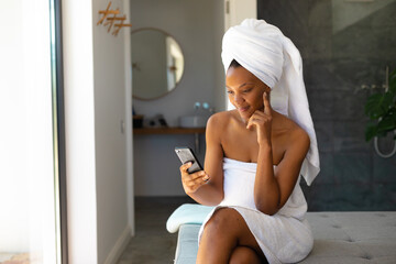 African american woman in a bathrobe using smartphone sitting on the bed at home - obrazy, fototapety, plakaty