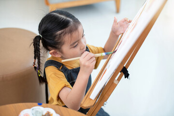 Close up, Asian little girl painting with water color in canvas standing on easel at home or school, little artist at work