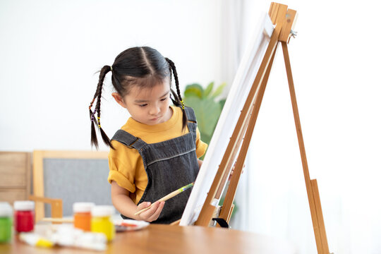 Asian little girl painting with water color in canvas standing on easel at home or school, little artist at work