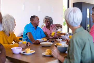 Multiracial seniors looking at happy woman serving juice to man at dining table in retirement home