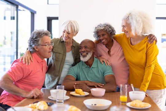 Multiracial senior friends with arms around enjoying by breakfast at dining table in nursing home
