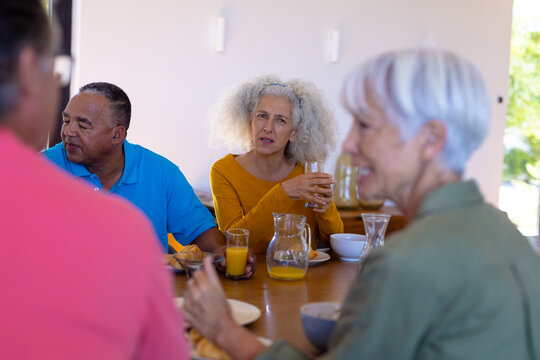 Caucasian senior woman holding juice looking at multiracial friends talking at dining table