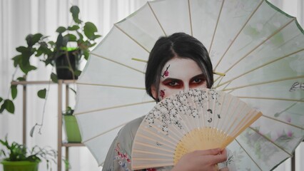A young beautiful Japanese-style girl stands with an umbrella and a fan in her hands and smiles cheerfully. Portrait of a stylish beautiful geisha under an umbrella with a sincere smile. Image of a