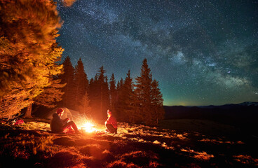 Pair of tourists spending evening time near bonfire on the middle of glade under open fullstar sky with Milky way in the mountains. Tent, trees, mountain peaks on the background. © anatoliy_gleb