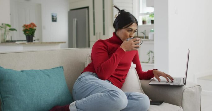 Video of happy biracial woman in hijab sitting on sofa with coffee and using laptop