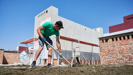 Full length of male worker shoveling sand-cement mix while working at construction site. Man in shorts preparing floor screed material while standing outdoors near concrete screed mixer machine.